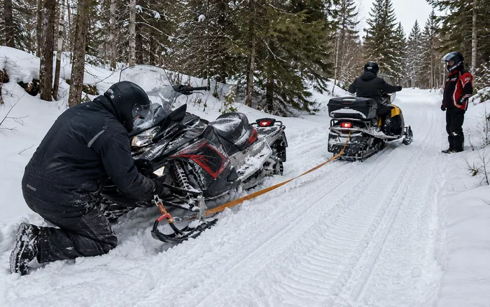 Snowmobile riders recovering a disabled sled on a snowy forest trail