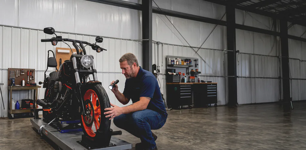 A Lakeview mechanic crouched with a flashlight, inspecting the front wheel and brake of a Harley-Davidson on a lift