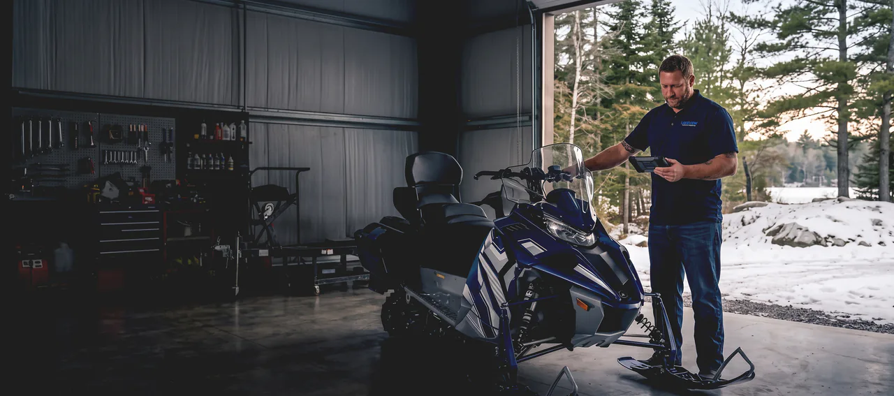 A Lakeview mechanic checking a sled on a diagnostic tablet in the open bay door, snow and pines behind him